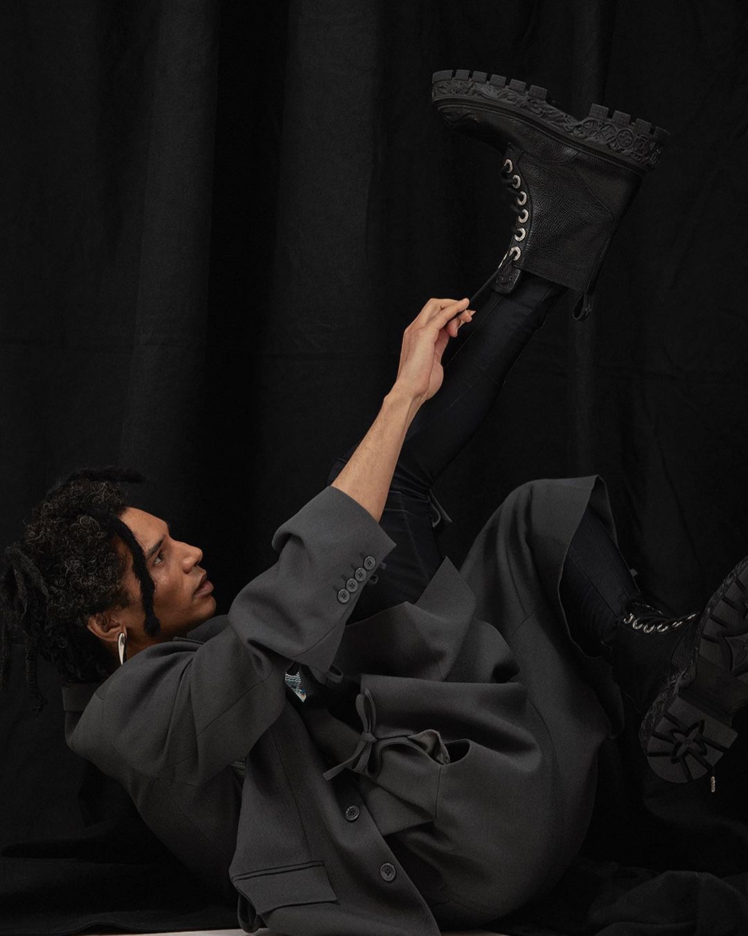 A person lying on a black background wearing a blazer, black pants, and a black boot with white laces being pulled up.