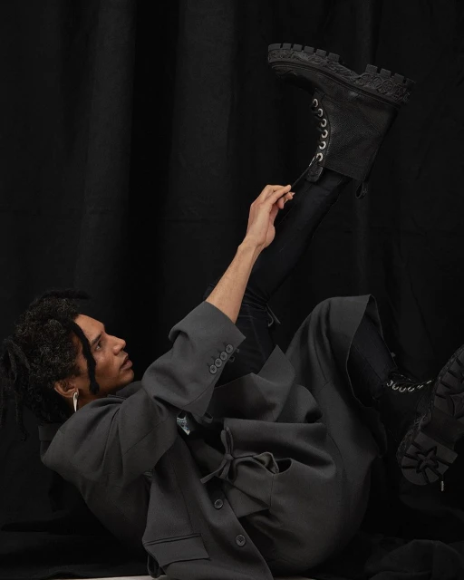 A person lying on a black background wearing a blazer, black pants, and a black boot with white laces being pulled up.