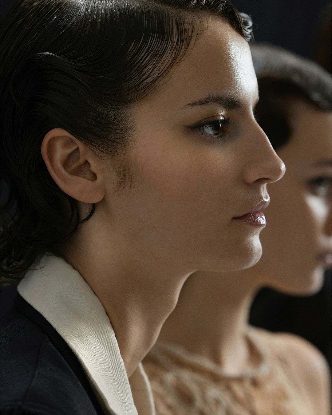 Close-up of two young women with dark, sleek hairstyles. One wears a dark jacket with a white collar; the other a textured, light brown top.