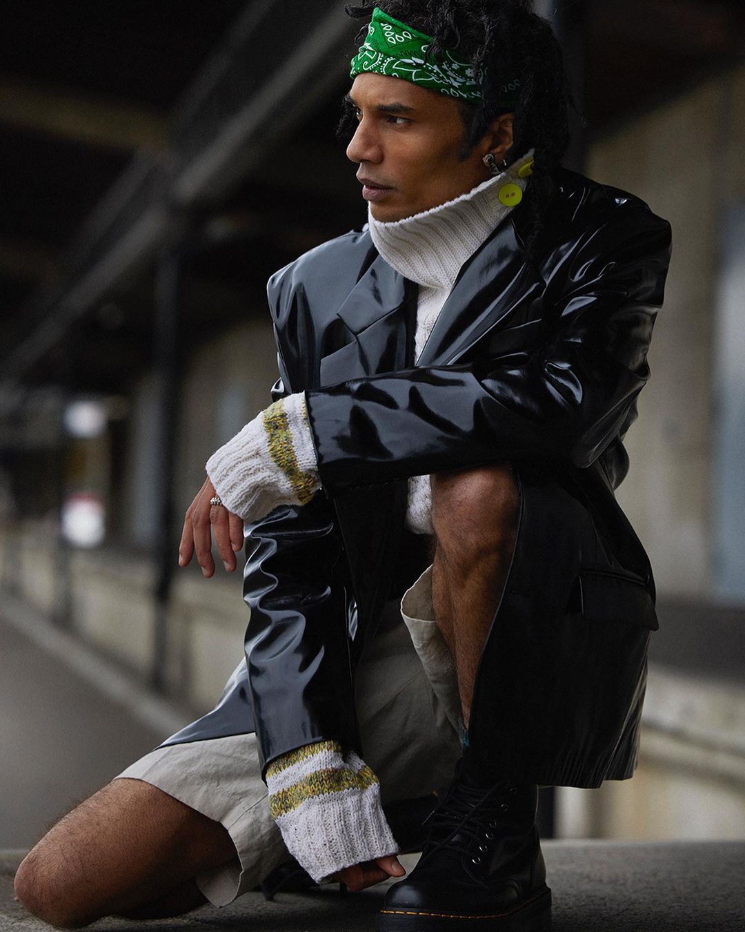 Man in a shiny black coat and green bandana kneels outside on concrete steps. He is wearing a white turtleneck and black boots.