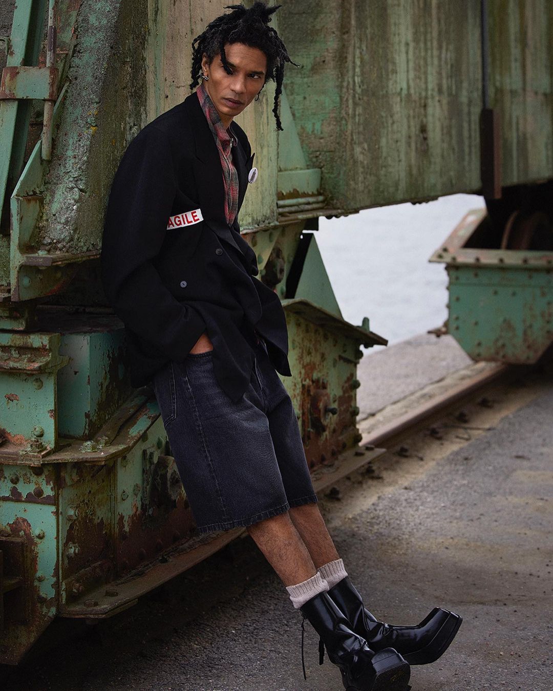 A person with dreadlocks in a black coat, black shorts, socks, and platform boots poses against an industrial background.