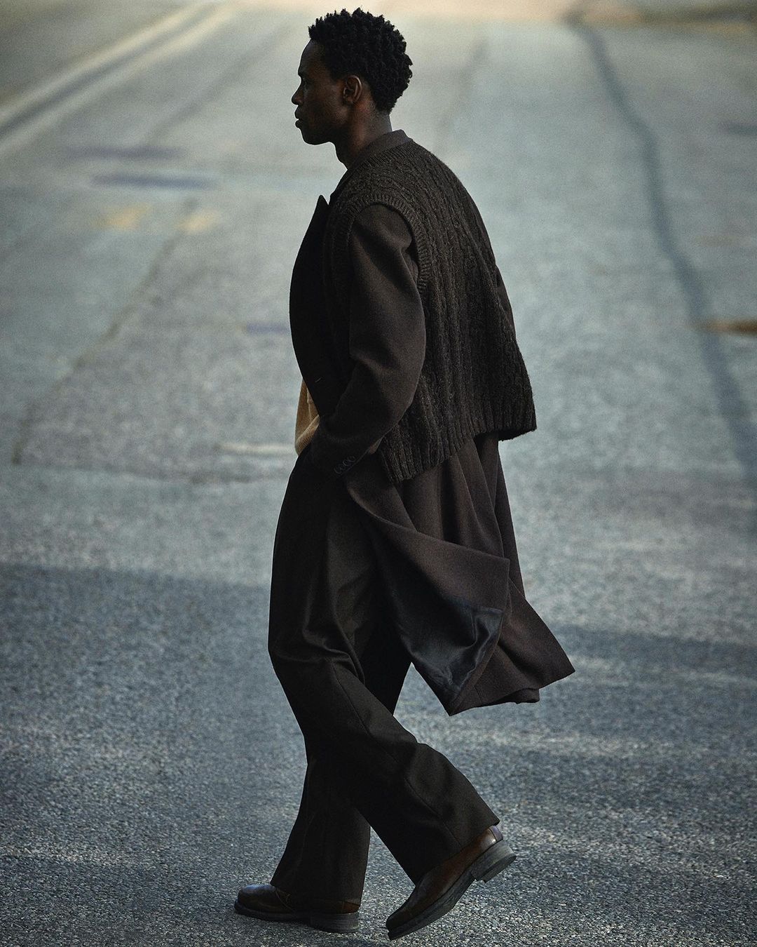 An adult man walks on an asphalt street wearing a dark brown knit vest, dark brown long coat, dark brown pants, and brown leather shoes.