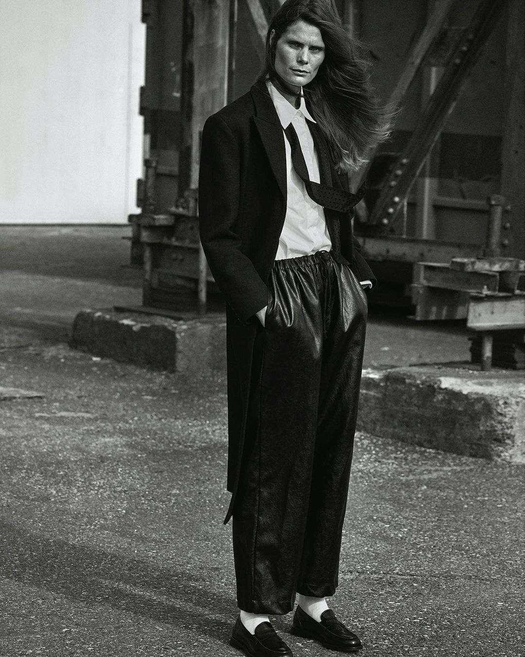 Monochrome portrait of a woman with long hair in a dark coat, white shirt, tie, and leather pants posing against an industrial backdrop.