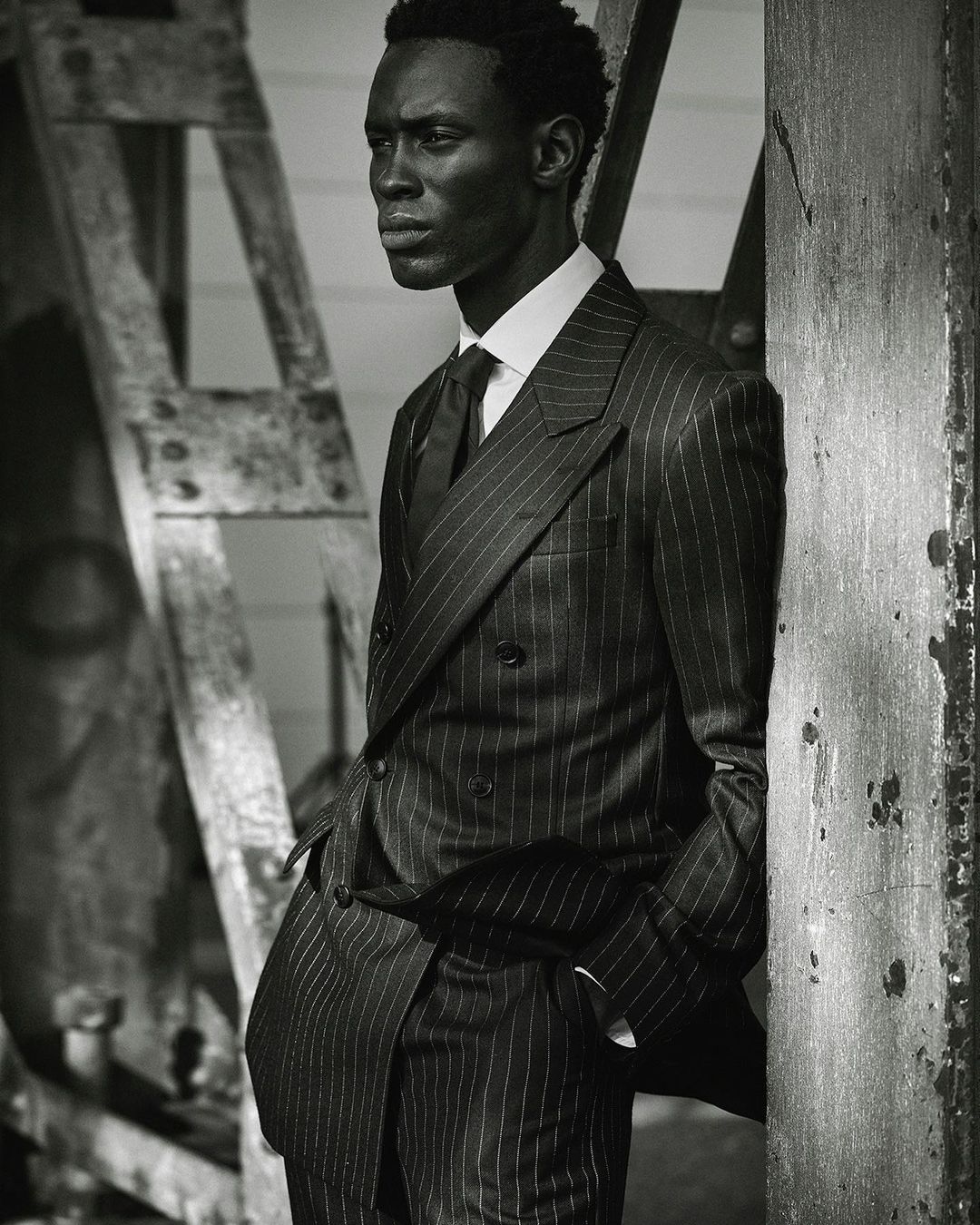 Portrait of an African American man in a pinstripe suit, standing against rustic wooden planks, captured in a black and white photograph.
