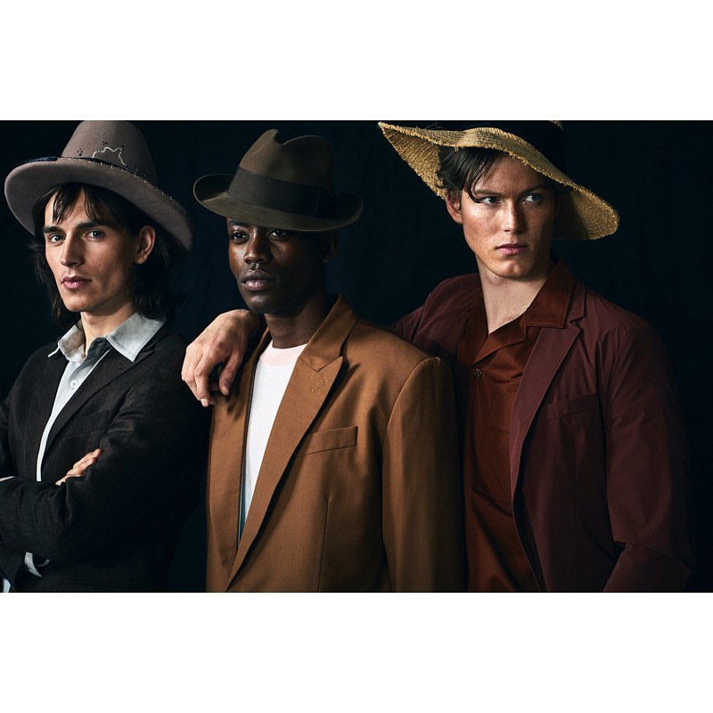 Three men in stylish blazers and hats stand close together against a dark background, showcasing a blend of textures and colors.