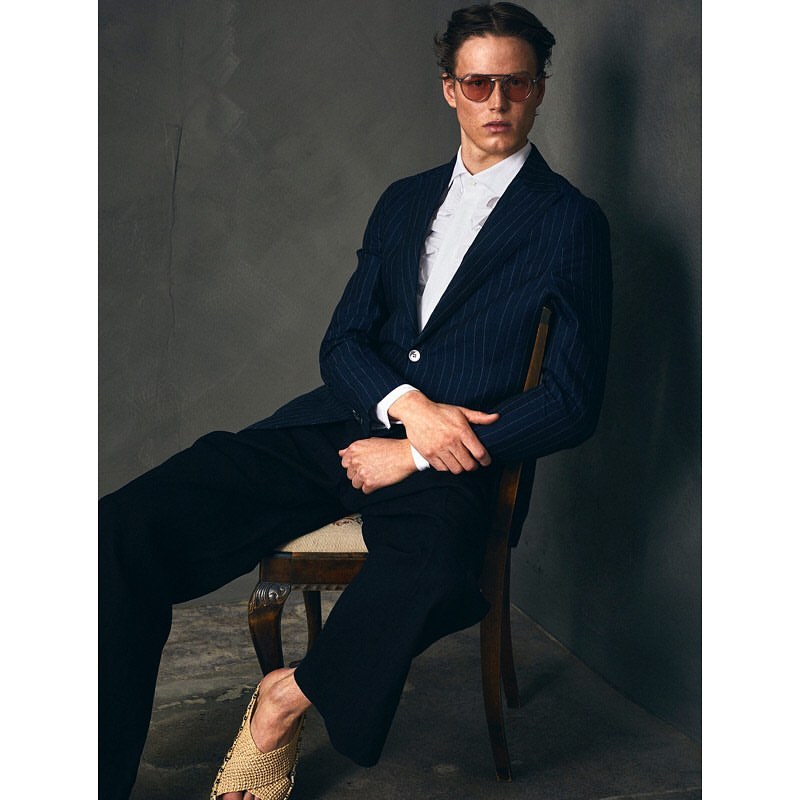 An adult male model sits on a chair wearing sunglasses, a striped blazer, a white button-down shirt, and dark trousers in a studio setting.