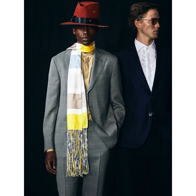 Two men are posing indoors in suits, with one wearing a red hat and a patterned scarf, set against a dark background.