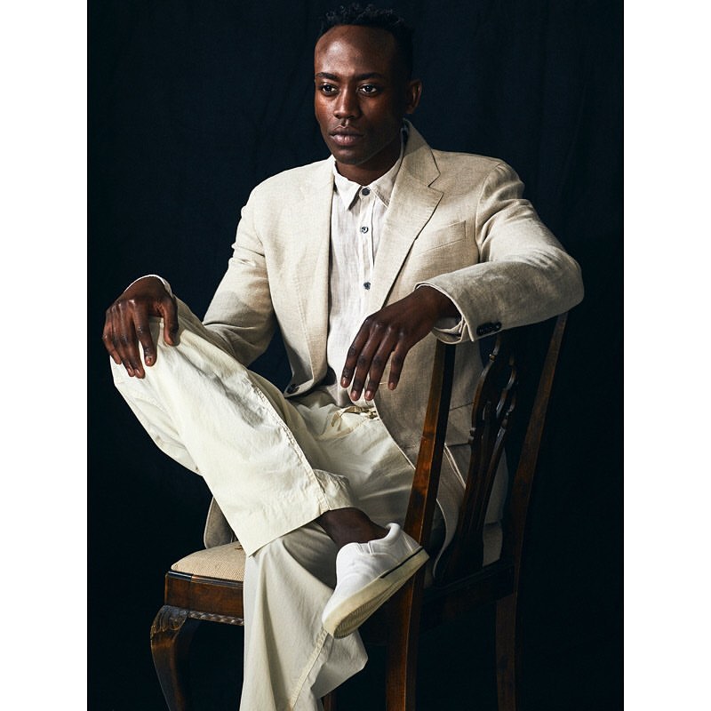 Adult man in a linen suit sits on a wooden chair in a studio with a black background, looking off camera with a neutral expression.