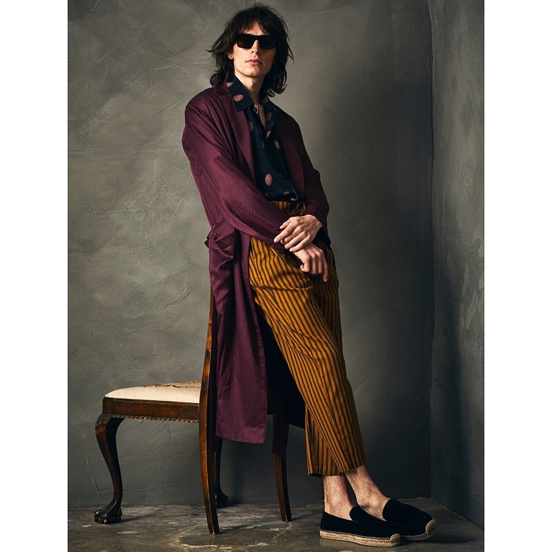 Man in a burgundy coat and striped pants leans against a chair in a studio, wearing sunglasses and dark loafers.