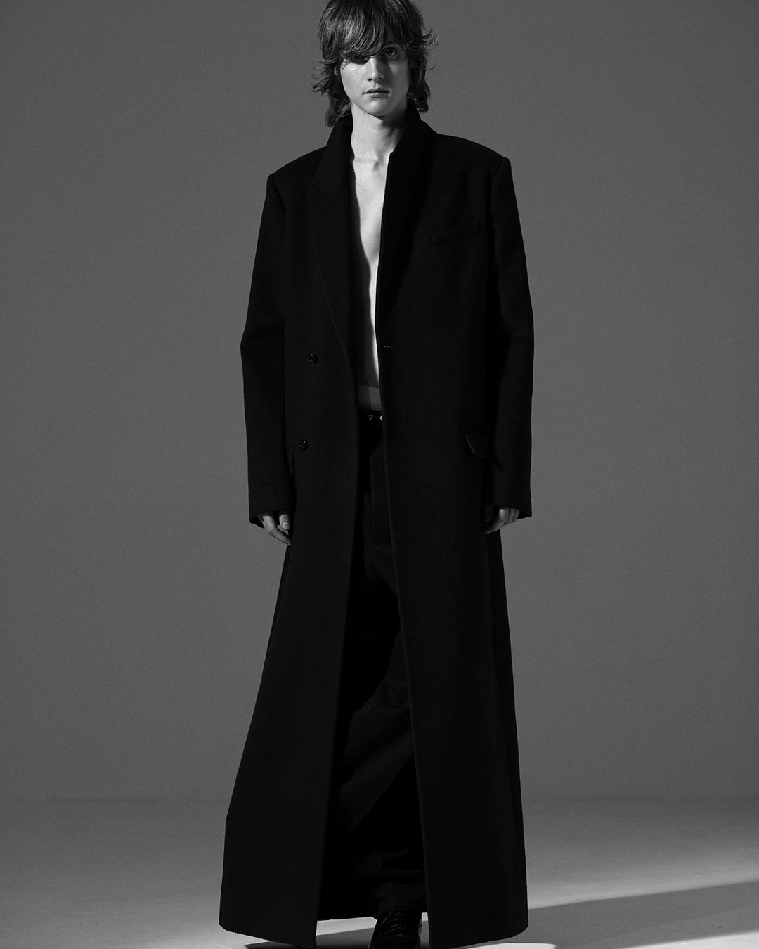Monochrome portrait of a slender adult male model in a long black coat, posing in a neutral gray studio setting, casting a dramatic shadow.