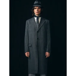 Man wearing a dark grey coat, fedora hat, and scarf standing against a plain black background in portrait framing.