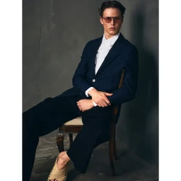 Man seated on a wooden chair wearing a pinstripe blazer, white ruffled shirt, dark trousers, and tinted sunglasses.