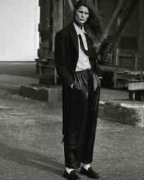 Model standing outdoors wearing a black coat, white shirt, black tie, and dark trousers photographed by Oscar Falk