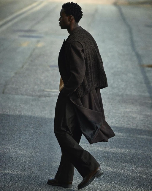 Man walking on an asphalt street wearing a dark brown overcoat and layered knit vest in Stockholm.
