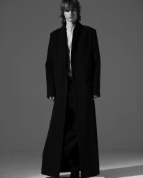 Monochrome portrait of a young male model wearing a long black coat over an open shirt, photographed against a neutral studio backdrop.