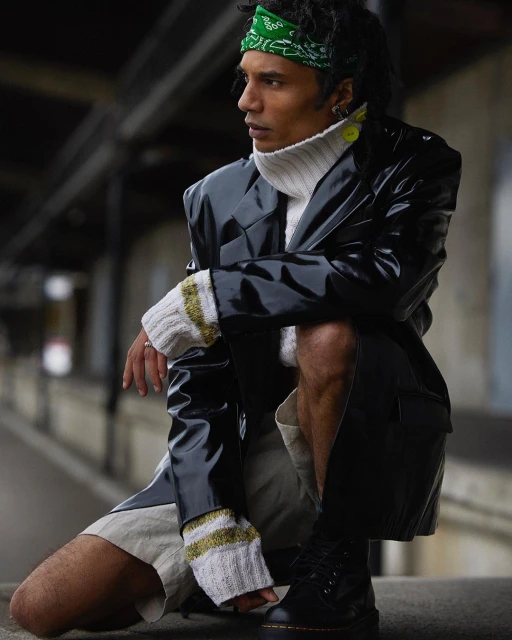 Man wearing a black patent jacket and white turtleneck crouching in an urban setting, photographed by Oscar Falk.