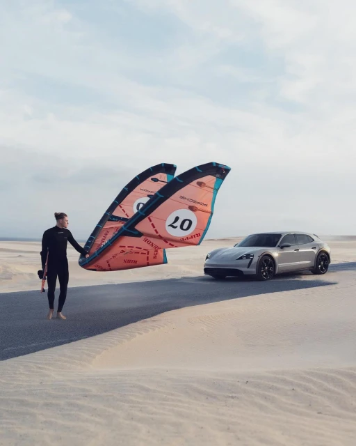 A kitesurfer holds two kites on a sand dune with a silver Porsche Taycan parked in the background under a clear blue sky.