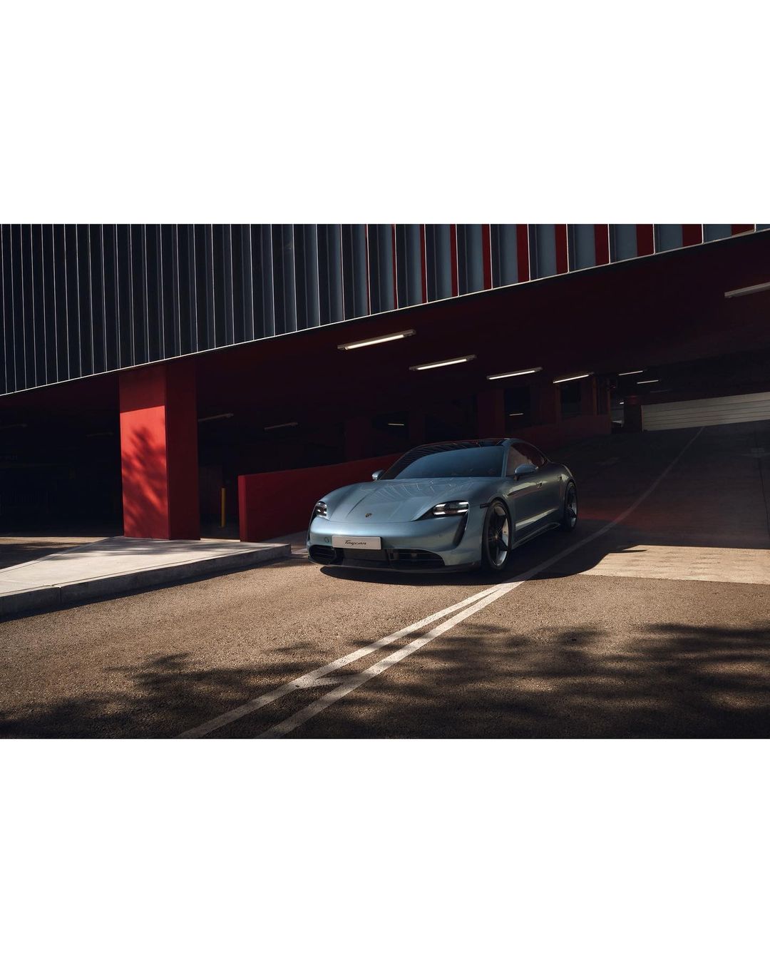 Silver Porsche Taycan parked under a modern building with grey slats and red pillars, showcasing automotive design and urban photography.