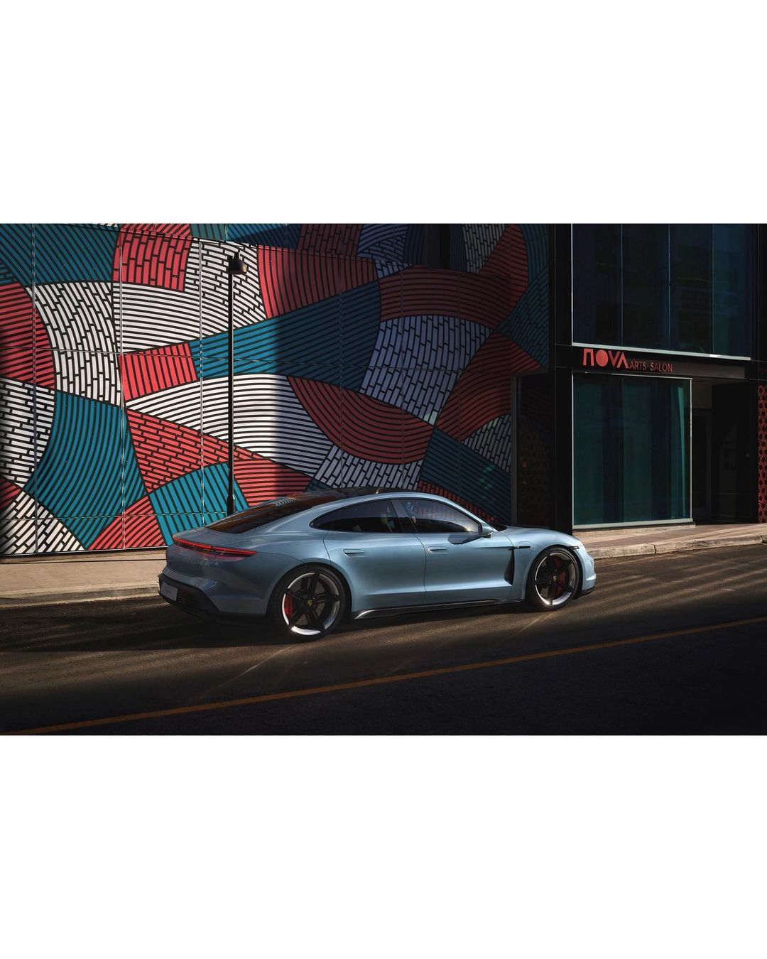 Light blue Porsche Taycan sedan parked on a city street, next to a building with colorful geometric mural and architecture. Car photography.
