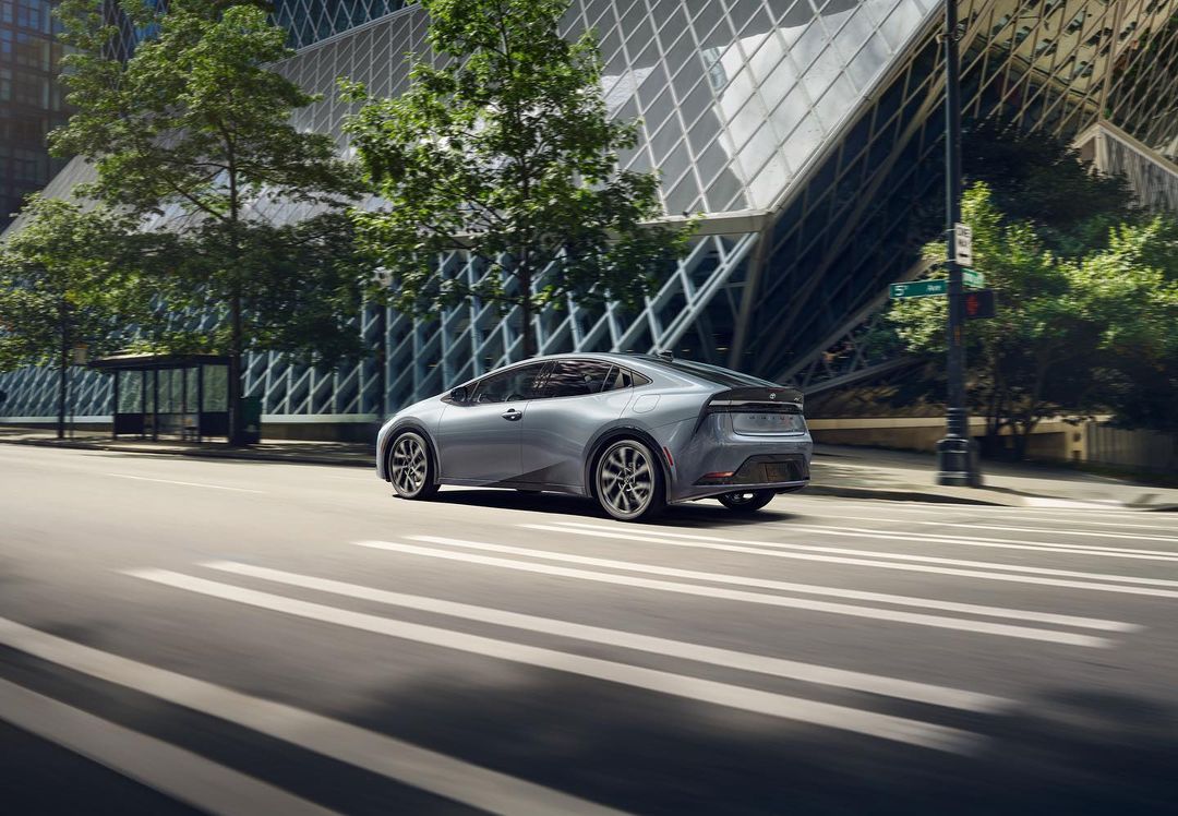 Silver car driving on a city street with a modern building and green trees in the background on a bright, sunny day.