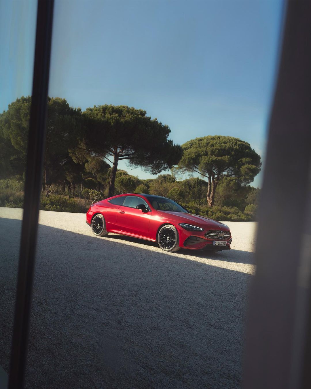 Red Mercedes coupe parked outdoors on gravel with green trees and a blue sky in the background on a sunny day.