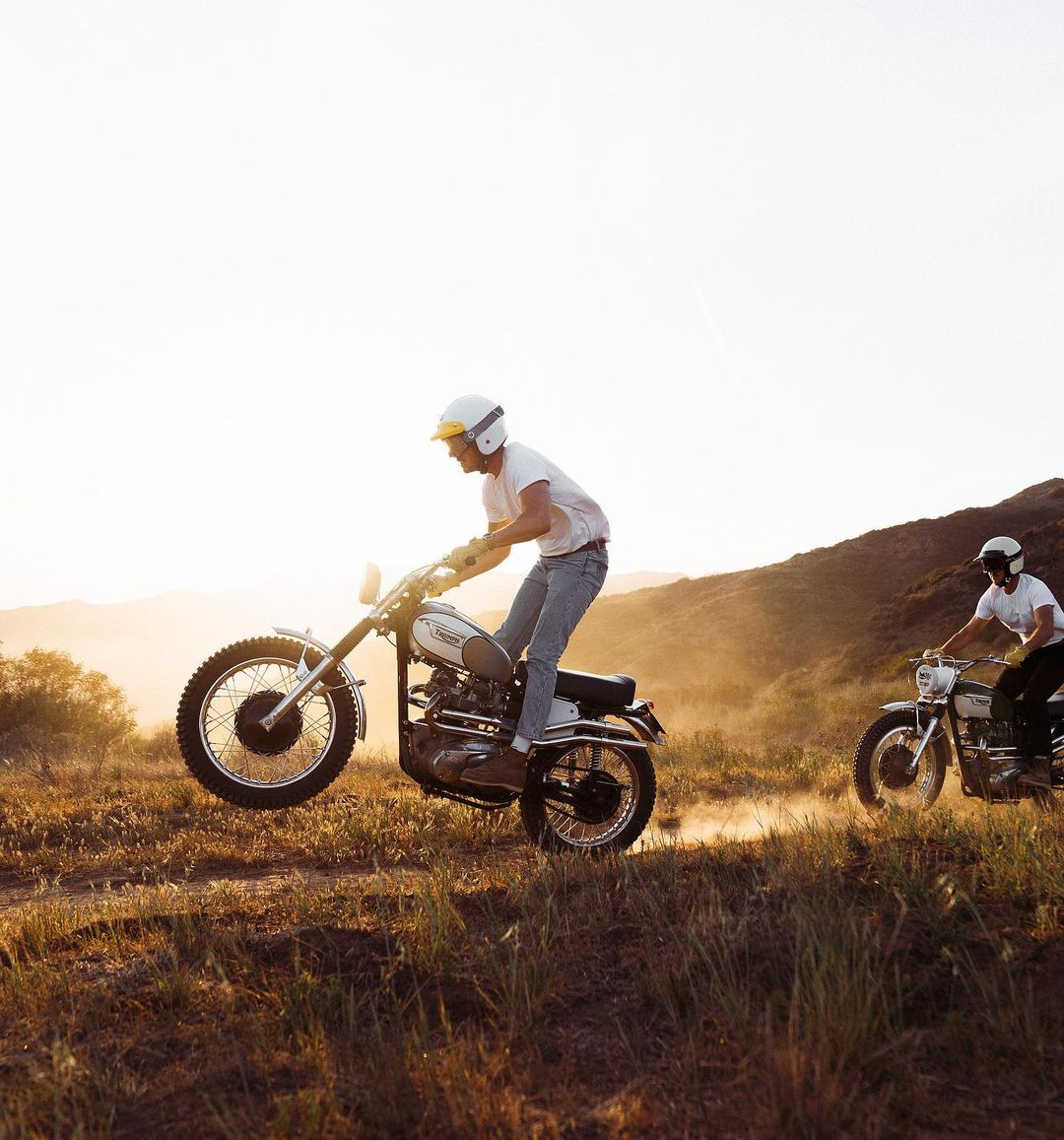 Two men riding vintage motorcycles on a grassy hill at sunset, showing a lifestyle image of outdoor recreation and classic bikes.