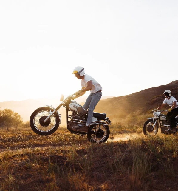 Two men riding vintage motorcycles on a grassy hill at sunset, showing a lifestyle image of outdoor recreation and classic bikes.