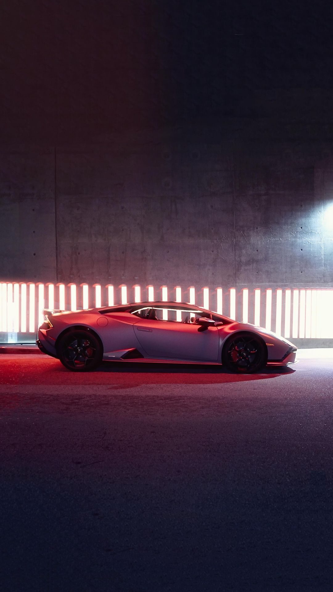 Sleek silver Lamborghini Huracan parked indoors with glowing red light panels behind it, side view, modern design and concrete floor.