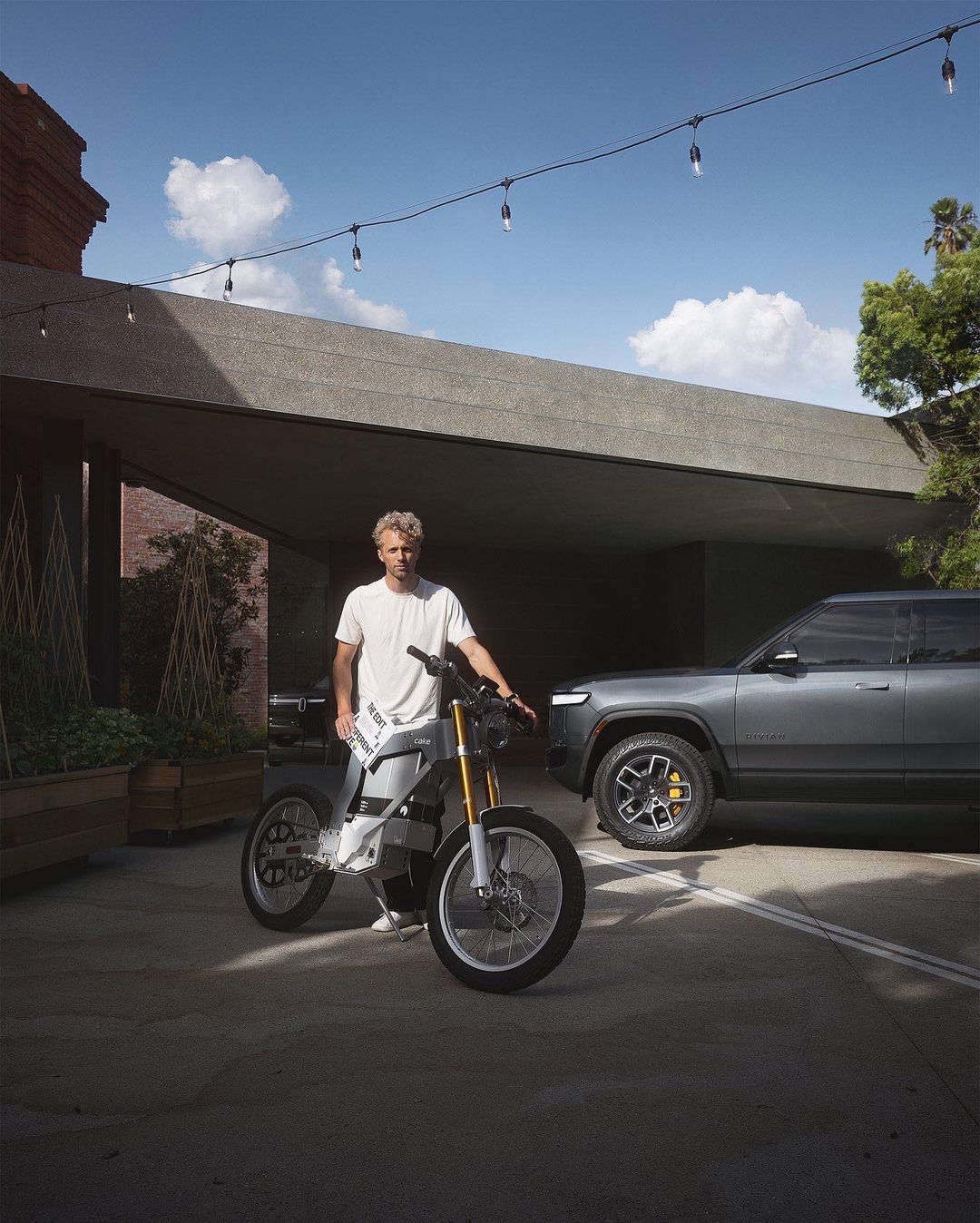 Man poses with an electric motorcycle near a parked Rivian SUV under bright sky with string lights; outdoor shot with modern architecture.