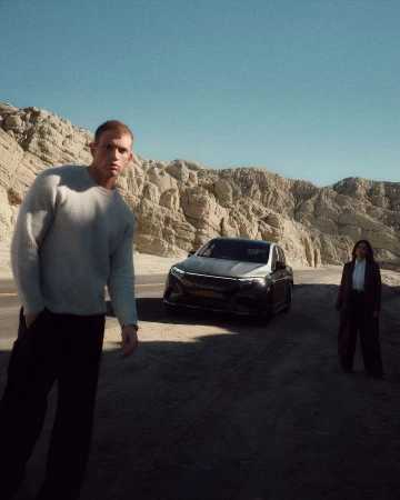 An electric car is parked on a desert road, flanked by a man in a sweater and a woman in a business suit, under a clear blue sky.
