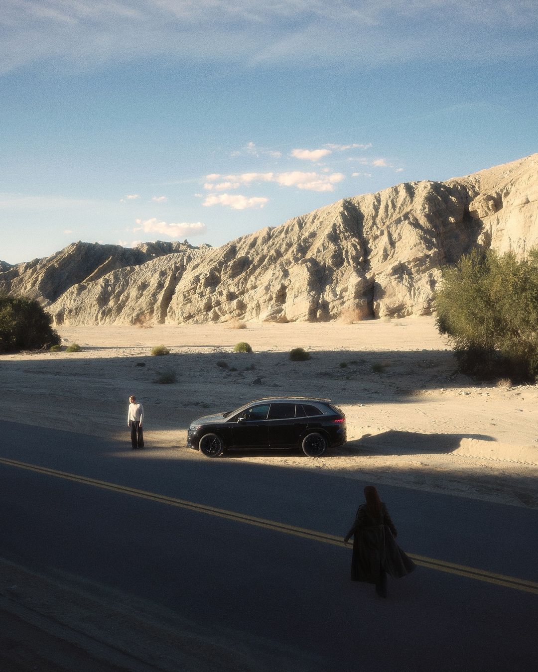 A black SUV is parked on a desert road with mountains, a male in white, and a female in black nearby on a sunny day.