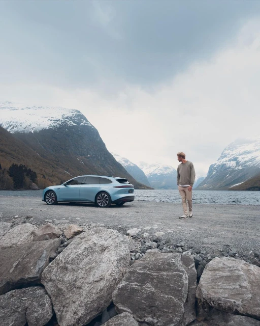 Light blue electric station wagon parked by a lake surrounded by snow-capped mountains, with an adult male standing nearby.