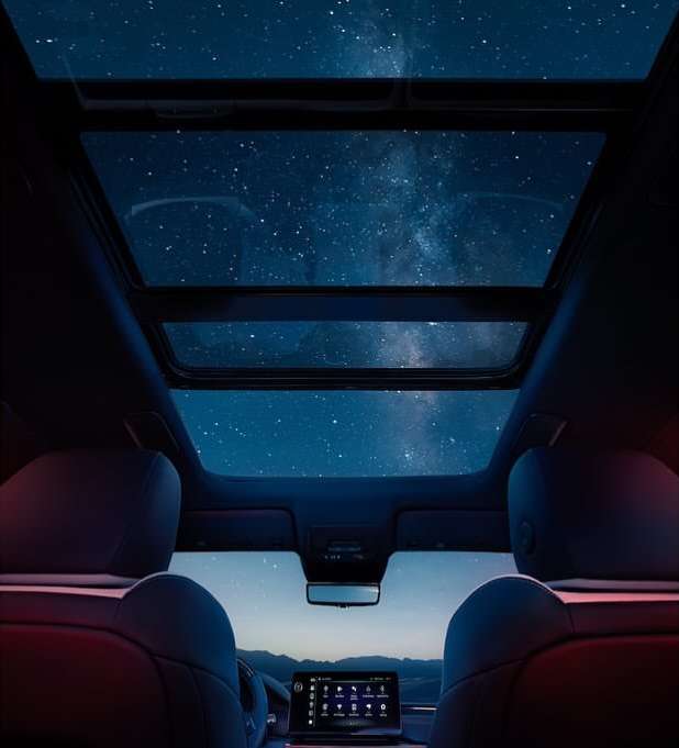 Interior of an automotive with a glass roof, showing the night sky with stars and a galaxy, seen from the backseat of the vehicle.