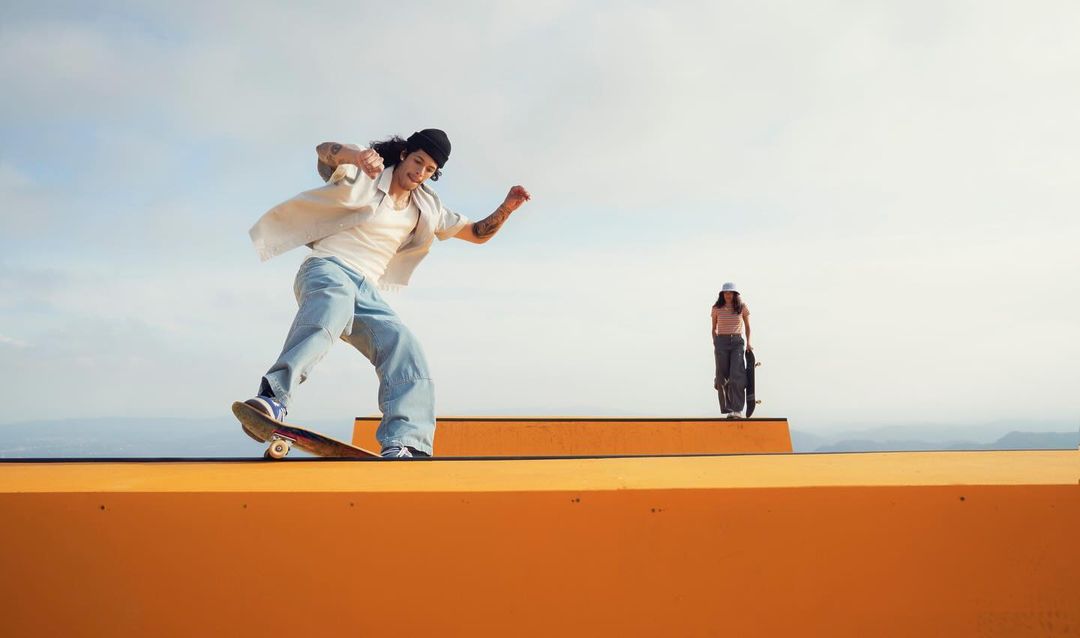 A skateboarder rides on a yellow ramp with another person standing in the background, set against a bright sky.