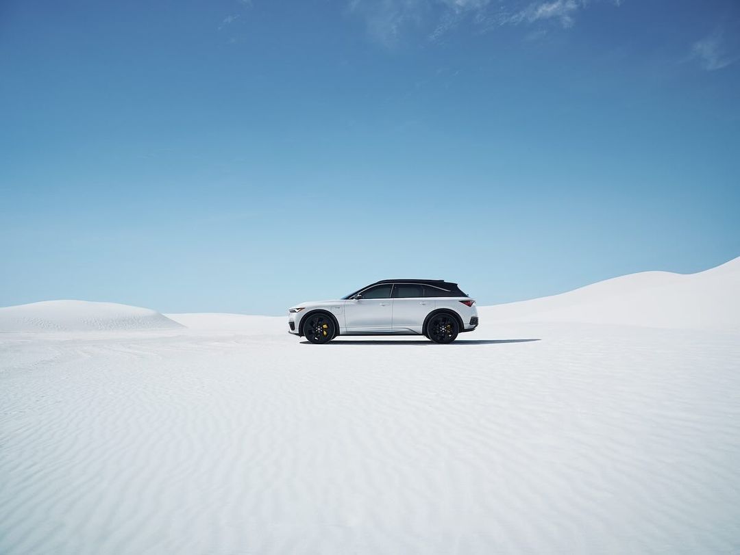 Silver SUV parked in a bright, stark desert landscape under a clear blue sky. The vehicle contrasts with the monochrome dunes.