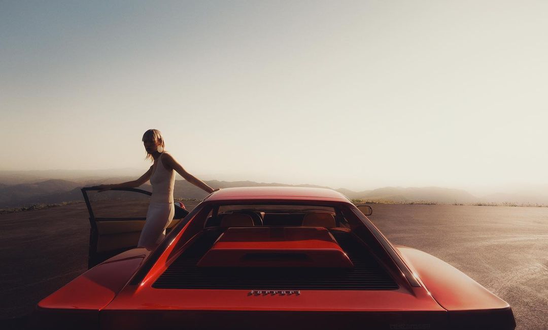 Woman in white dress by red Ferrari on a hilltop at sunset. The car's taillights and rear are visible, overlooking a mountain landscape.