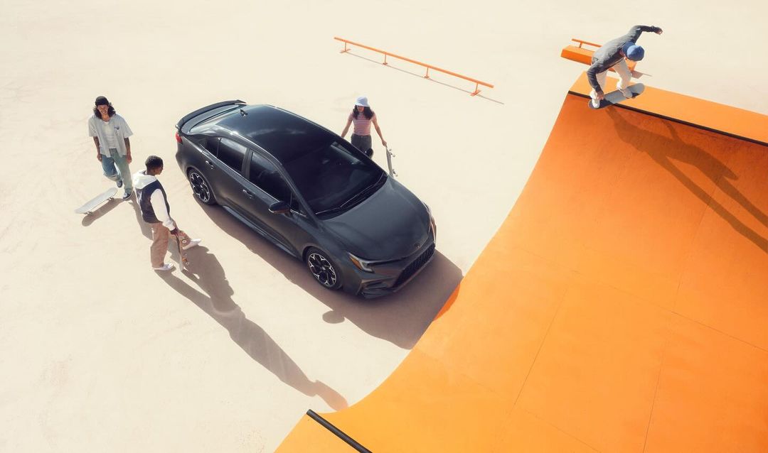 Aerial view captures a sleek grey hatchback car parked near a skatepark ramp with skateboarders and a tan surface under bright light.
