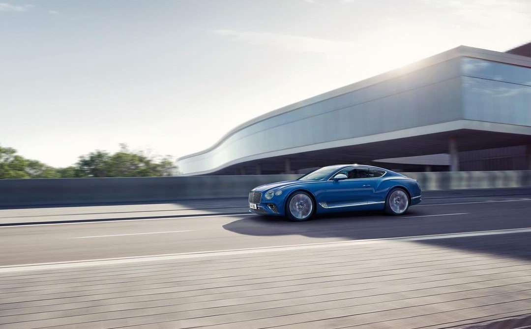 Blue Bentley coupe speeding along a road with a modern building, trees, and sky in the background, captured on a bright day.