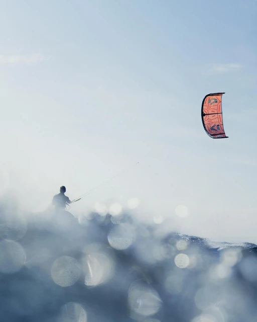 A person kite surfing with a red and black kite in the air above a wave of water on a sunny day with a clear blue sky.
