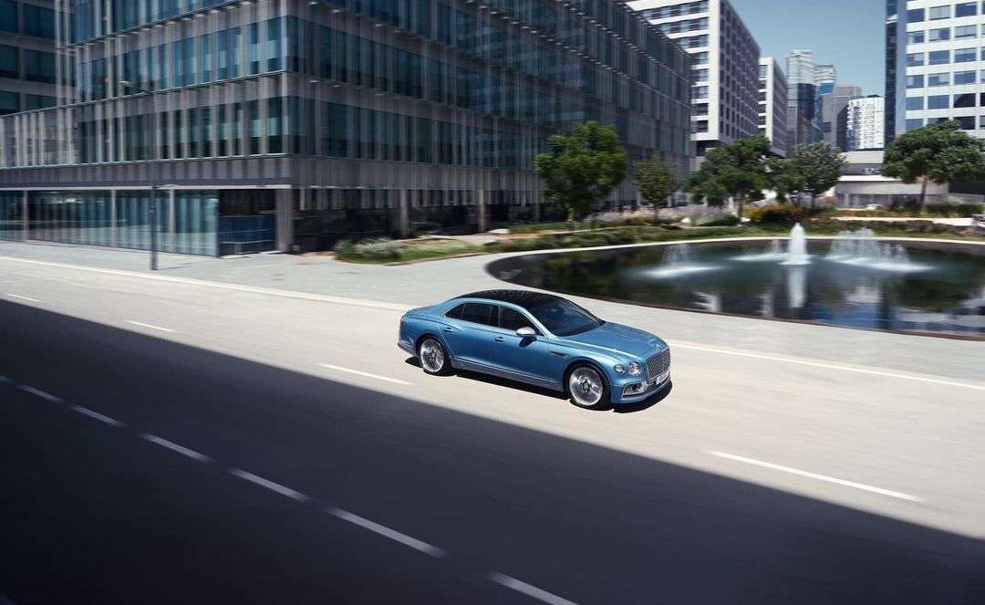 A blue Bentley sedan drives along a city street on a sunny day, with tall buildings and a water feature in the background.