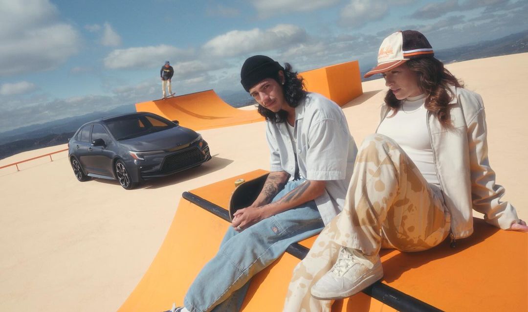 Automotive ad with a gray car positioned near skateboard ramps, featuring two young adults and one skateboarder in a desert environment.