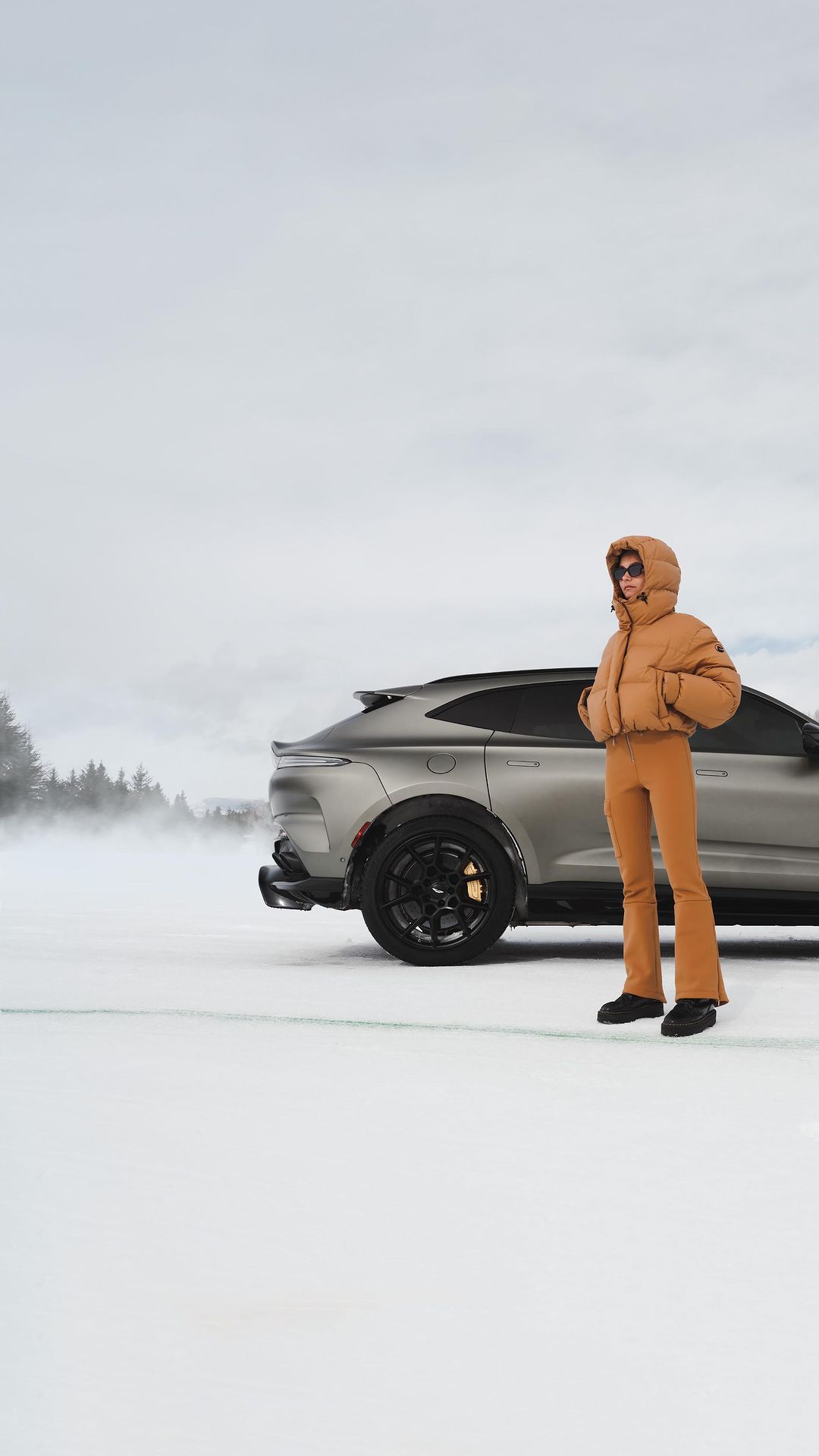 Woman in a brown puffer jacket standing on a snowy field next to a dark grey luxury SUV