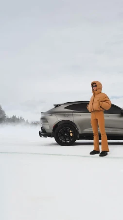 Woman in a brown puffer jacket standing on a snowy field next to a dark grey luxury SUV