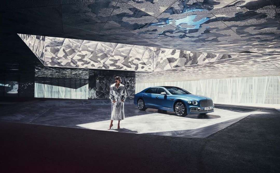 A luxury blue Bentley sedan is parked indoors while a woman in a long, shiny coat stands nearby in a spacious area.
