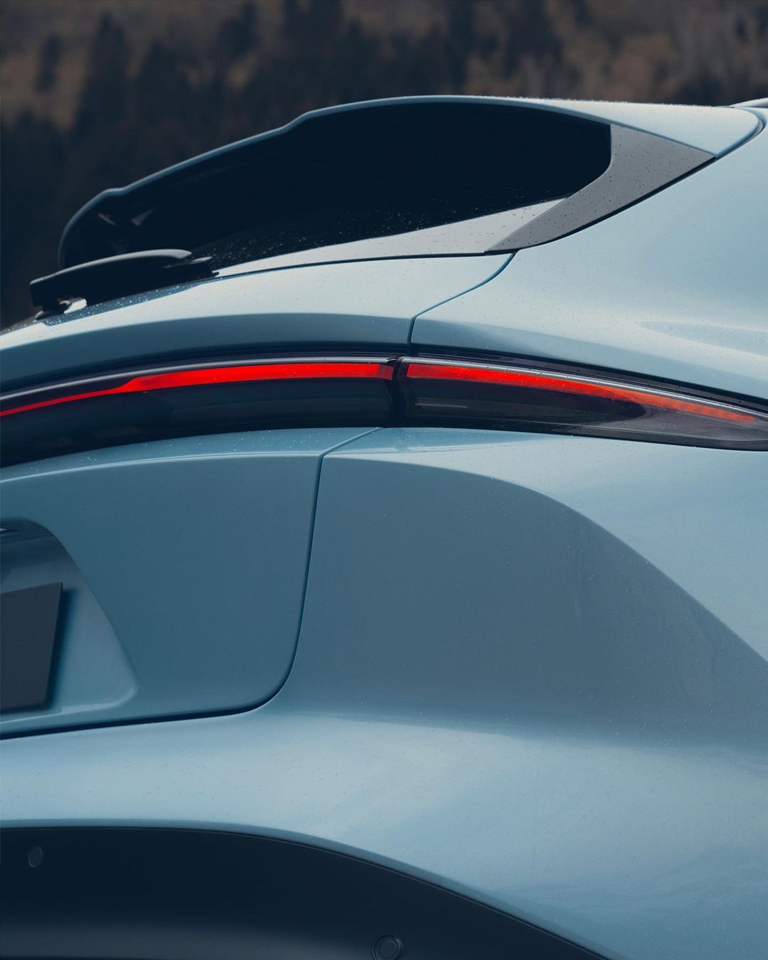 Close-up shot of the rear of a light blue crossover SUV with red tail light against a blurred, dark woodland background.