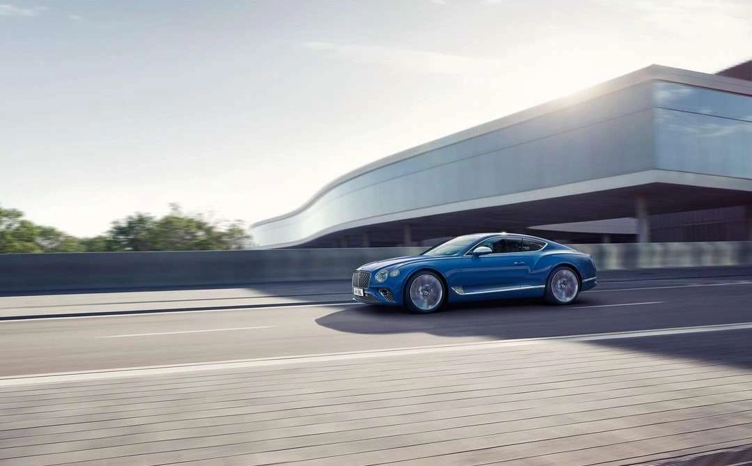 A blue sports car driving on a coastal road past a modern glass building under bright daylight.