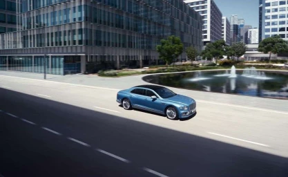 A light blue luxury sedan driving on an open city street past modern office buildings and a circular fountain.