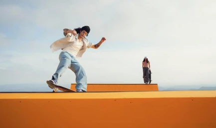 Skater man performing a trick on an orange ramp with a woman holding a skateboard in the background under an overcast sky.