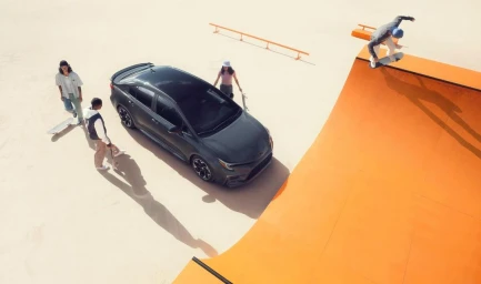 High angle shot of a dark sedan parked near a bright orange ramp with skaters outdoors in sunlight