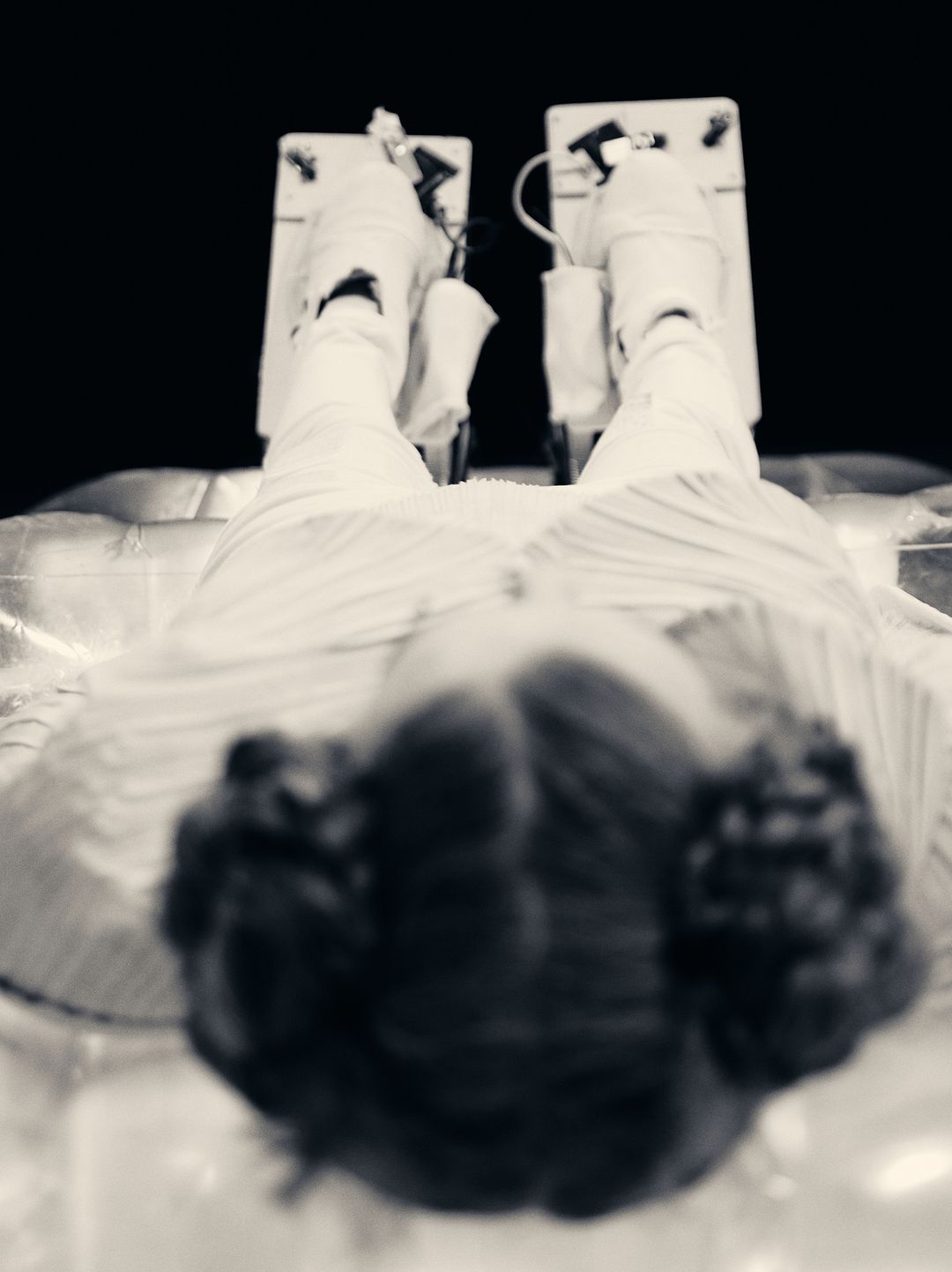 Black and white eye-level shot of a woman lying down with legs elevated on cushioned supports, with dark hair and striped clothing.